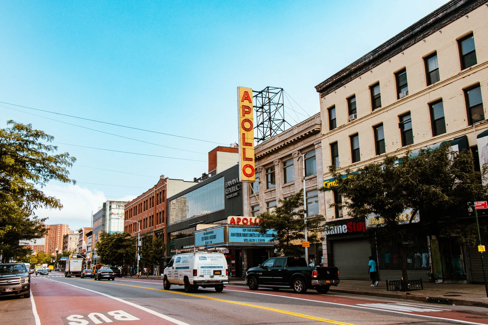 L'alto insegna verticale e il marquee dell'Apollo Theater sulla 125th Street ad Harlem, accanto a una corsia preferenziale rossa per autobus e a edifici in mattoni sotto un cielo azzurro